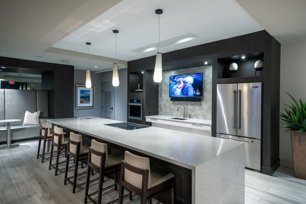 Modern kitchen with island seating, pendant lights, built-in TV, stainless fridge, and light wood floors.