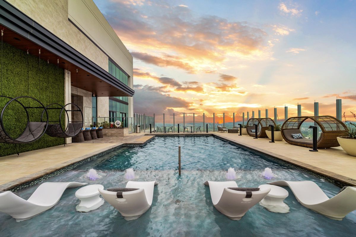 Rooftop pool with loungers at sunset, surrounded by lounge chairs and a glass barrier overlooking a cityscape.