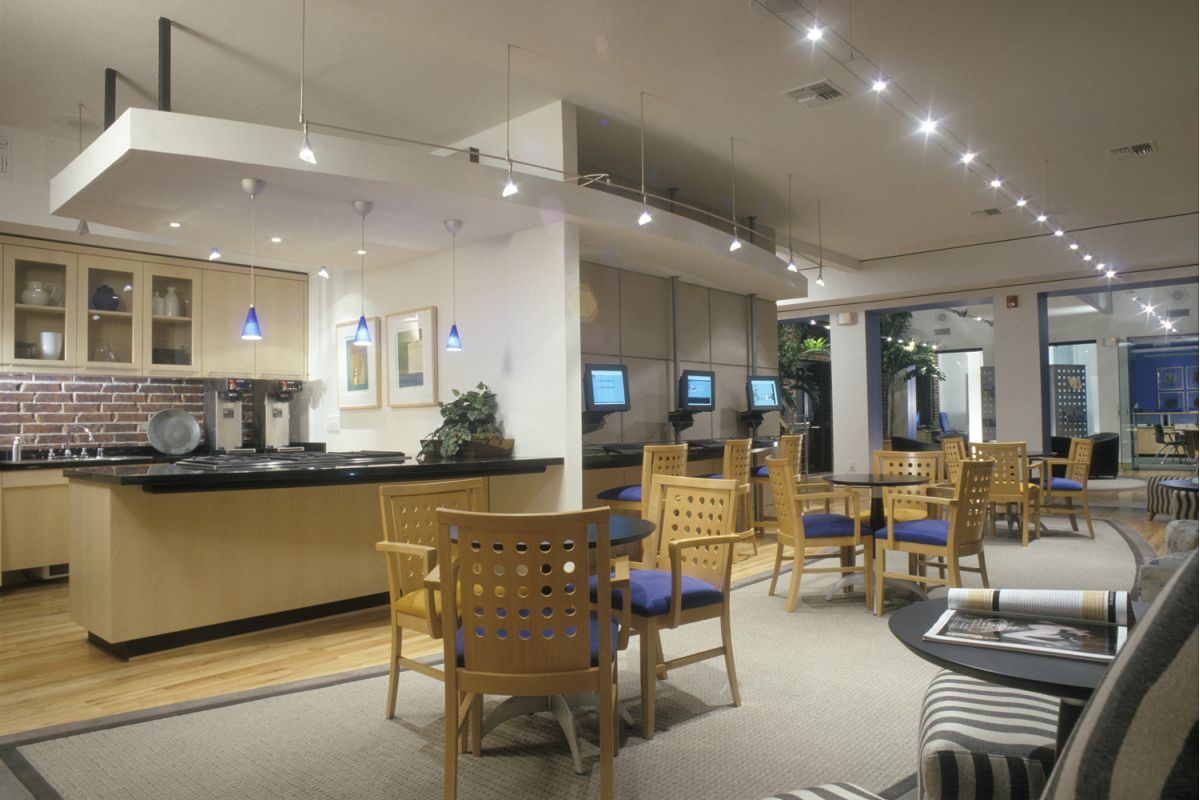 Modern lounge with wooden chairs, tables, computers, and a kitchenette with coffee machines and blue pendant lights.