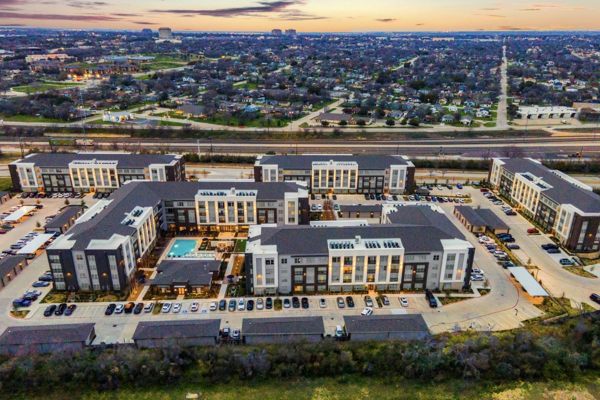 Aerial view of a large apartment complex at sunset with a visible pool and surrounding cityscape.