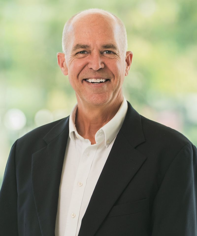Smiling older man in a dark blazer and white shirt standing outdoors with a blurred green background.