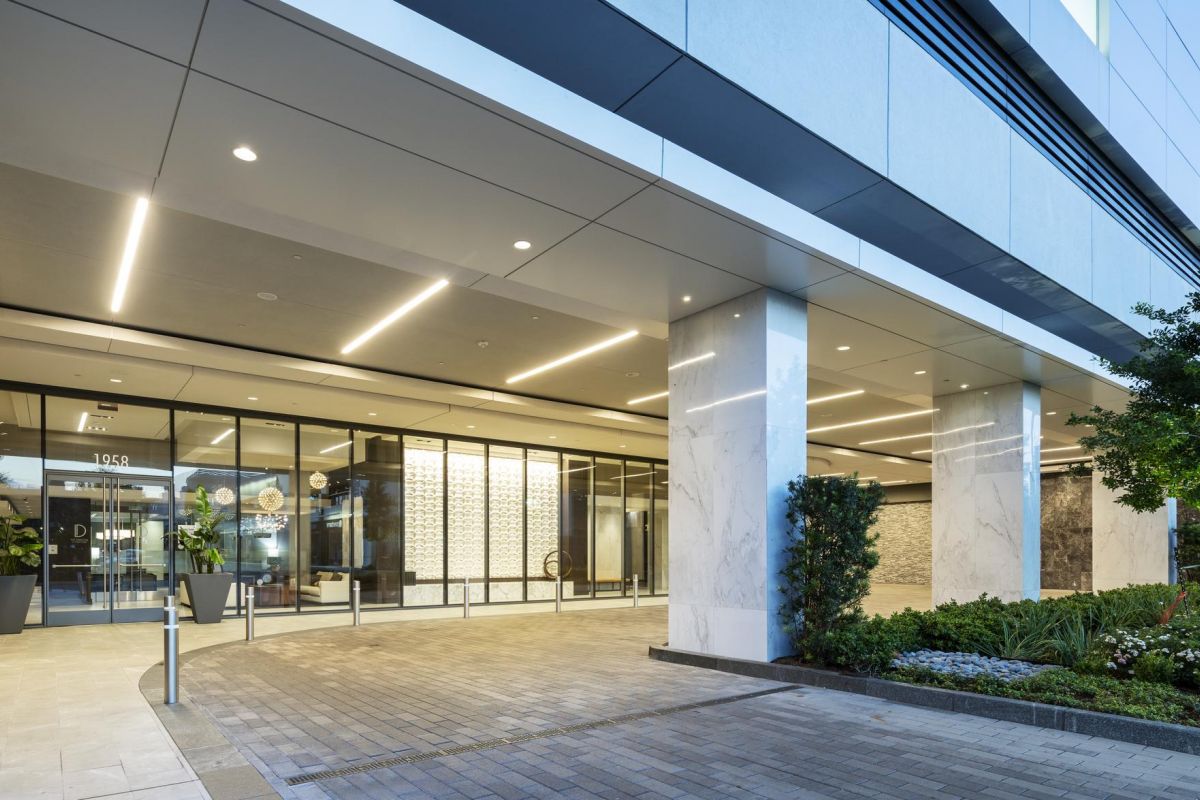 Modern office building entrance with glass doors, illuminated by sleek overhead lighting, surrounded by plants.