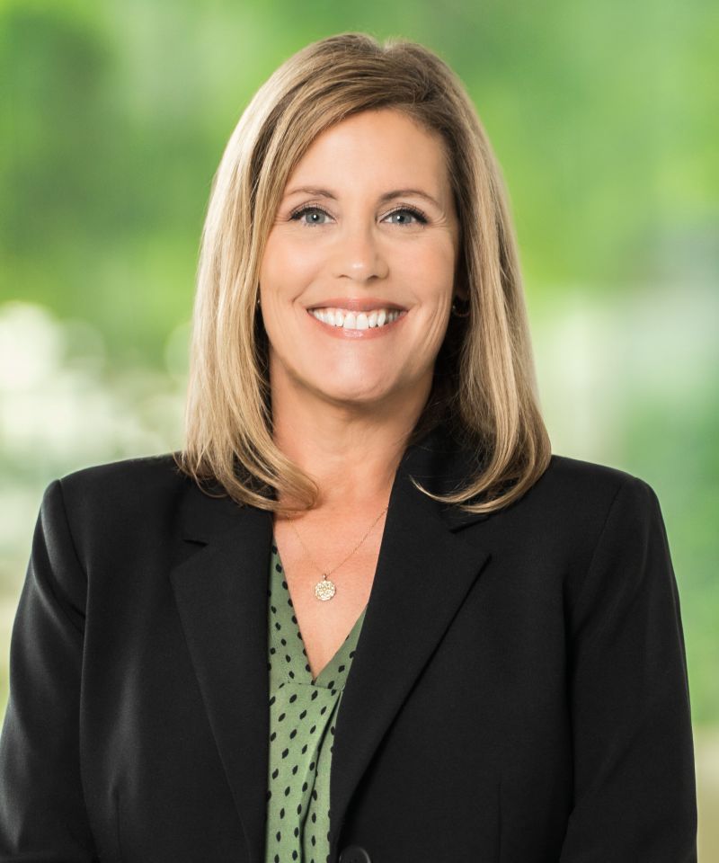 Smiling woman with shoulder-length blonde hair in a black blazer and green blouse, standing outdoors.