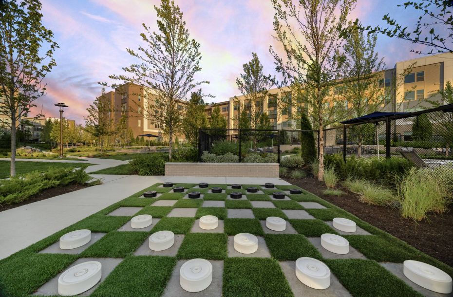 Hanover Company, Hanover Colony Place Outdoor checkerboard with large pieces on a grassy area, surrounded by trees and modern buildings at sunset.