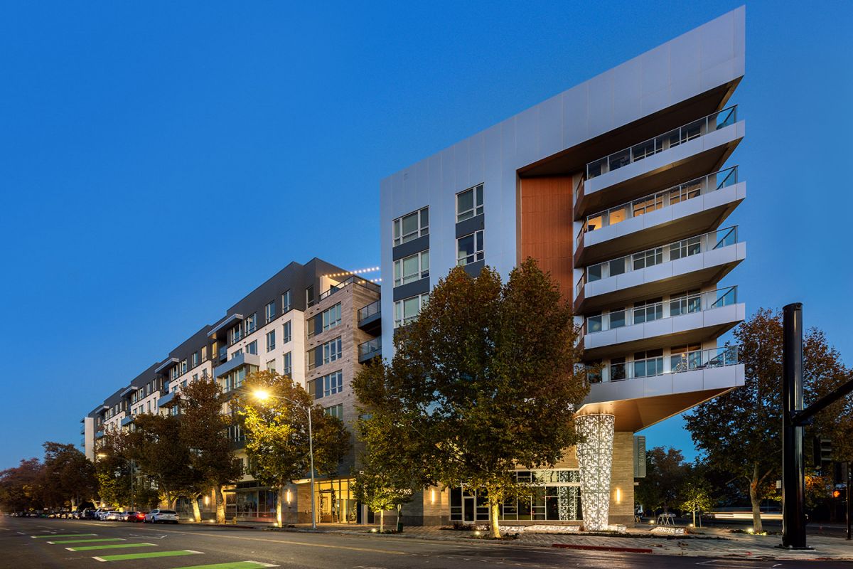 Modern multi-story Hanover Diridon apartment building with balconies, lit by streetlights at dusk, trees lining the sidewalk.