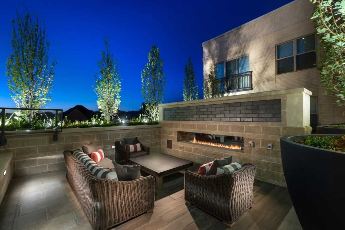 Outdoor patio at night with wicker sofas, a table, and a modern built-in fireplace, surrounded by trees.