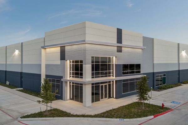 Modern industrial warehouse building with large windows, surrounded by pavement and a few small trees.