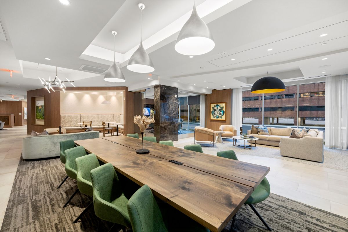 Modern lounge with a long wooden table, green chairs, sofas, large windows, and pendant lighting.