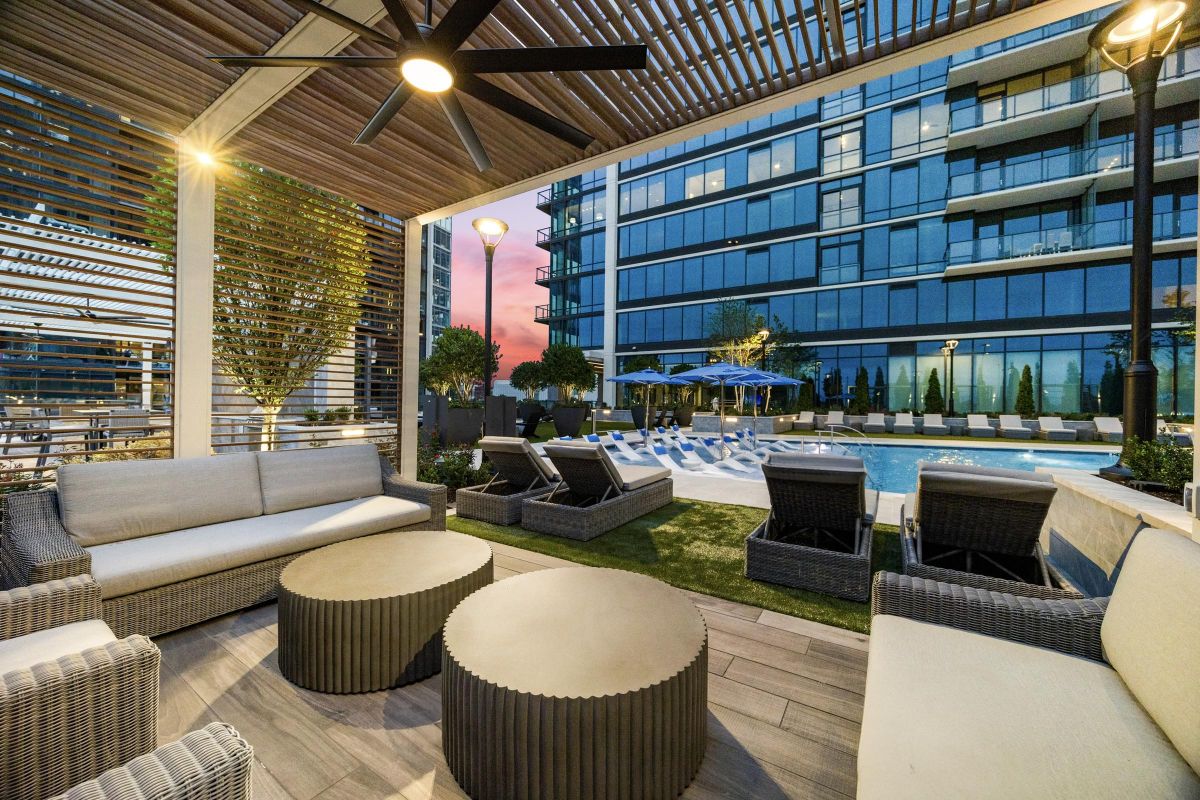 Modern outdoor lounge with sofas and tables overlooking a pool and apartment building at sunset.