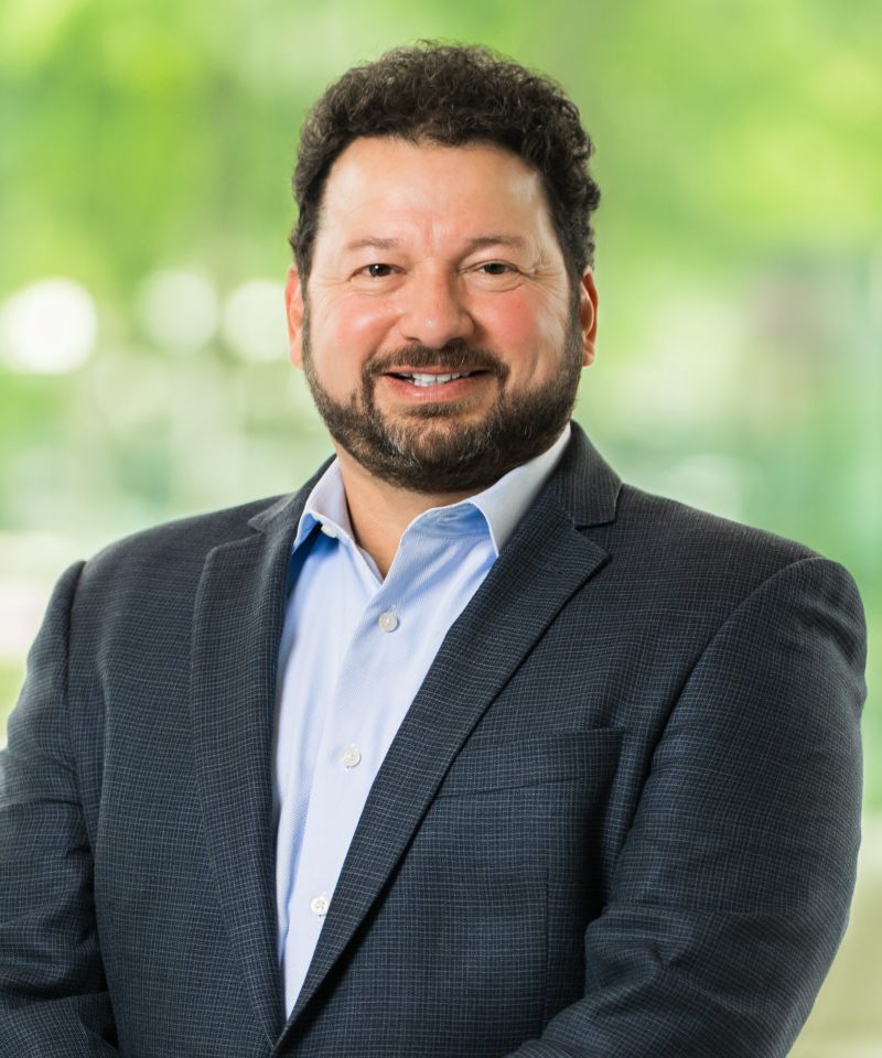 Man with curly hair and beard wearing a blue suit jacket and light shirt, smiling in front of blurred green background.