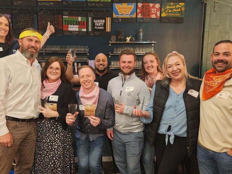 Group of smiling adults with drink glasses, some wearing bandanas, posing together in a bar or brewery setting.