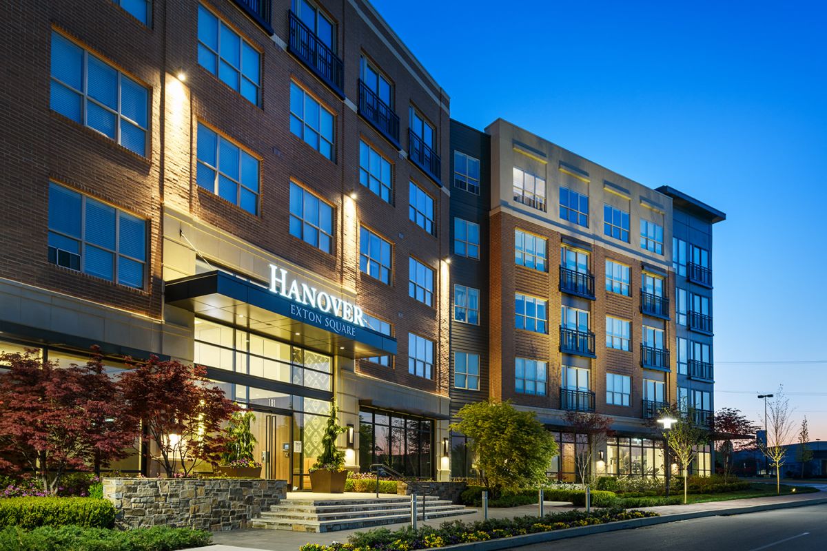 Modern apartment building with large windows and bright exterior lights at dusk, sign reads "Hanover Exton Square.