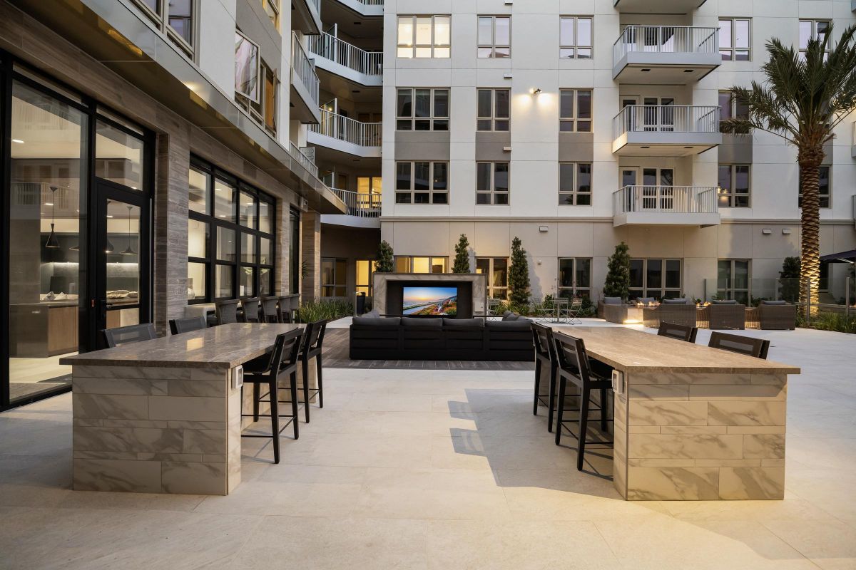 Hanover Laguna Niguel Modern outdoor patio with communal tables, chairs, and a large TV, surrounded by apartment buildings and palm tree.