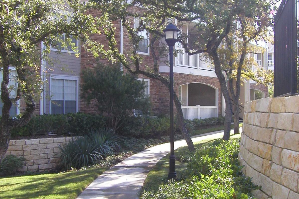 Curved sidewalk lined with greenery and trees, beside an apartment building with balconies and stone walls.