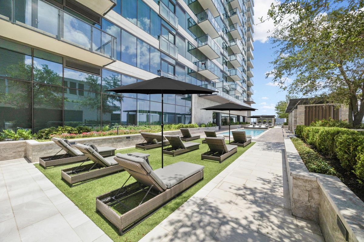 Outdoor lounge area with wicker chaise lounges and umbrellas near a modern building, with a pool in the background.
