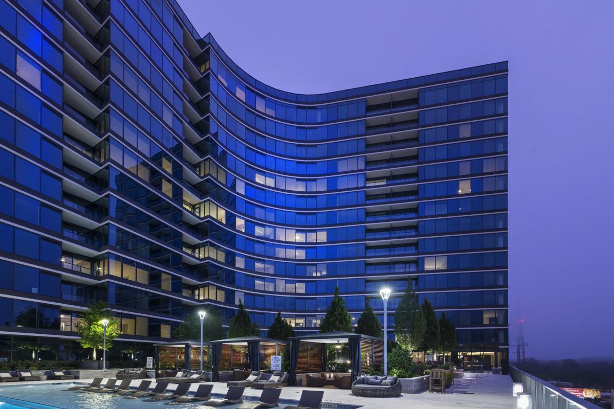 Modern high-rise building with a pool and lounge chairs, illuminated at dusk.