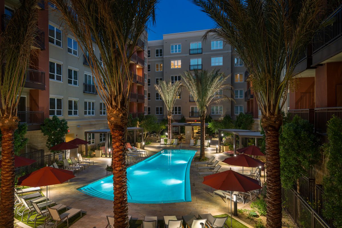 Evening view of a well-lit apartment pool area with palm trees, lounge chairs, and red umbrellas.