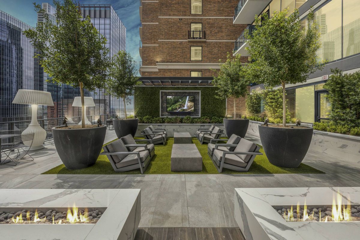 Hanover Company, Hanover Republic Square Modern rooftop patio with seating, planters, fire pits, and a large screen, surrounded by city buildings at dusk.