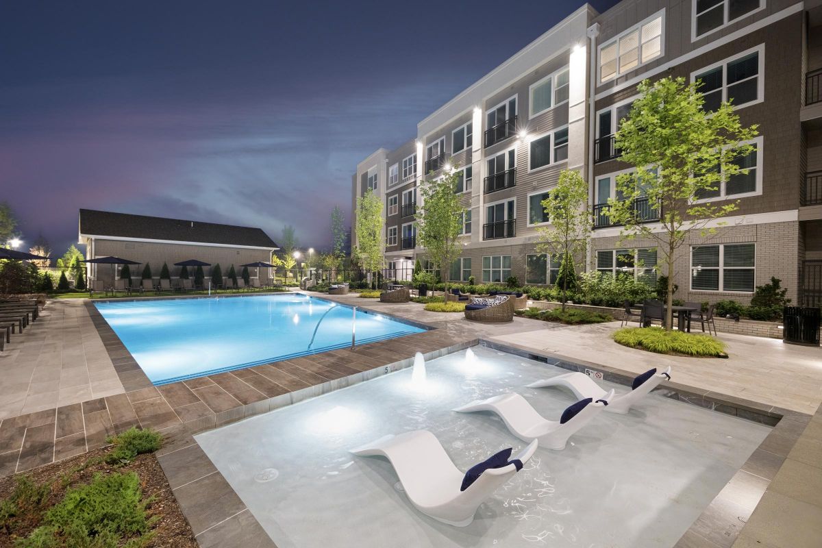 Modern apartment complex with a lit swimming pool, loungers in a shallow pool, and trees at dusk.