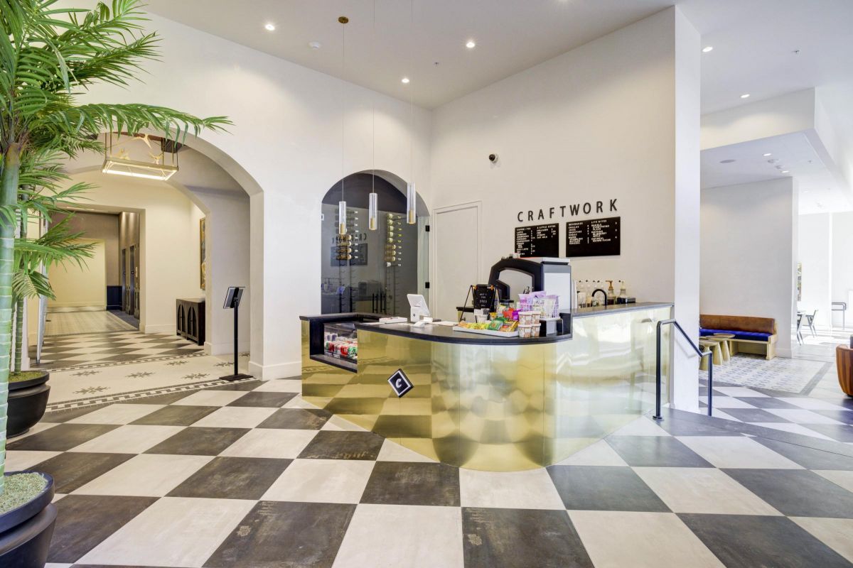 Modern lobby with a gold reception desk, checkered floor, and greenery. Sign reads "CRAFTWORK." Bright lighting.