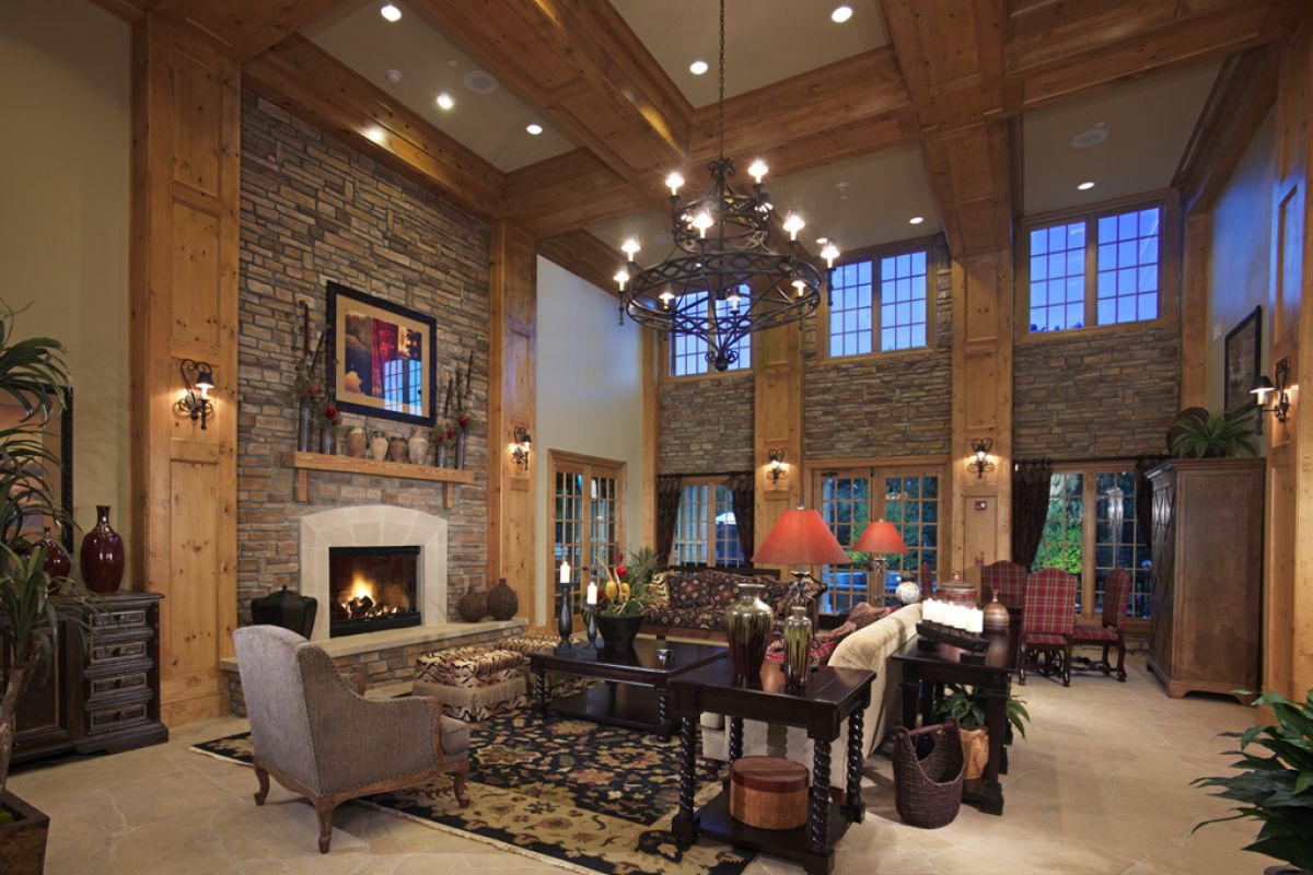 Spacious living room with stone fireplace, wooden beams, large windows, and cozy seating under warm lighting.