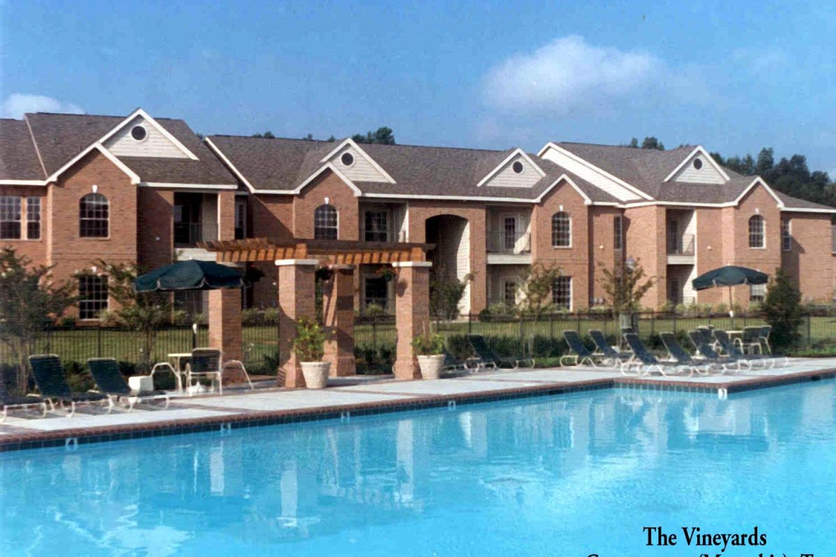 Outdoor swimming pool with lounge chairs and umbrellas in front of a two-story brick apartment building.
