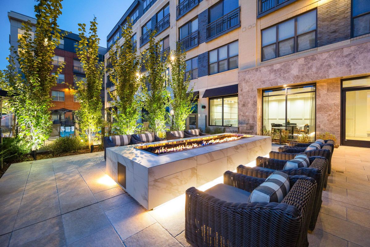 Modern outdoor patio with a long fire pit, wicker seating, and lit trees near an apartment building at dusk.