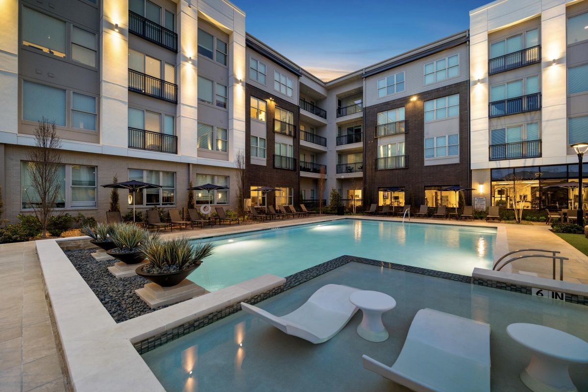 Modern apartment complex with illuminated windows and a pool featuring submerged lounge chairs, at dusk.
