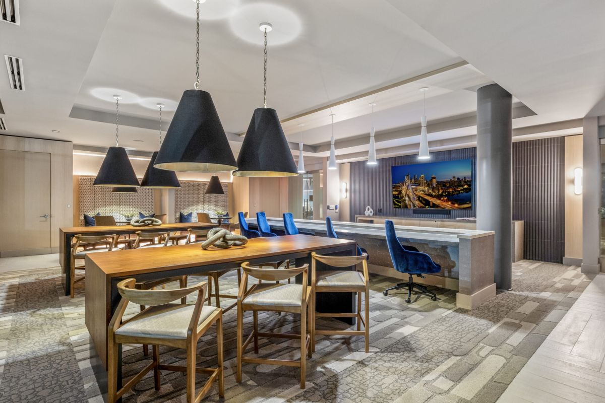 Modern Stoneham lounge with wooden tables, pendant lights, blue chairs, and a large TV on the wall.