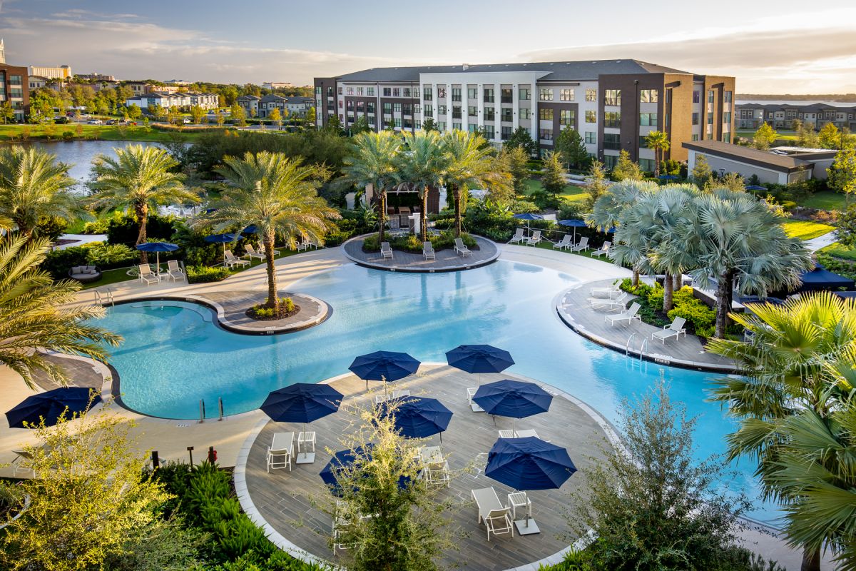 Resort-style pool with palm trees, lounge chairs, and blue umbrellas near apartment buildings at sunset.