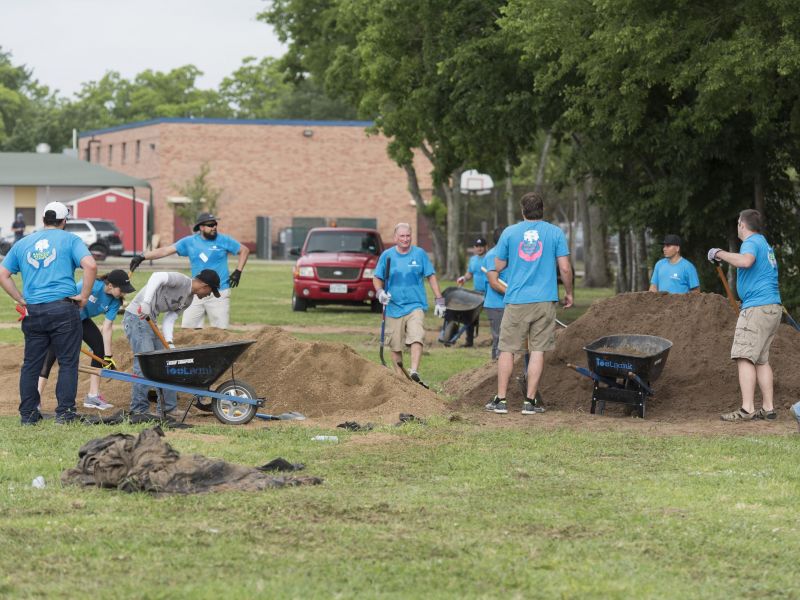 People in blue shirts move sand with wheelbarrows and shovels in a grassy outdoor area with trees.