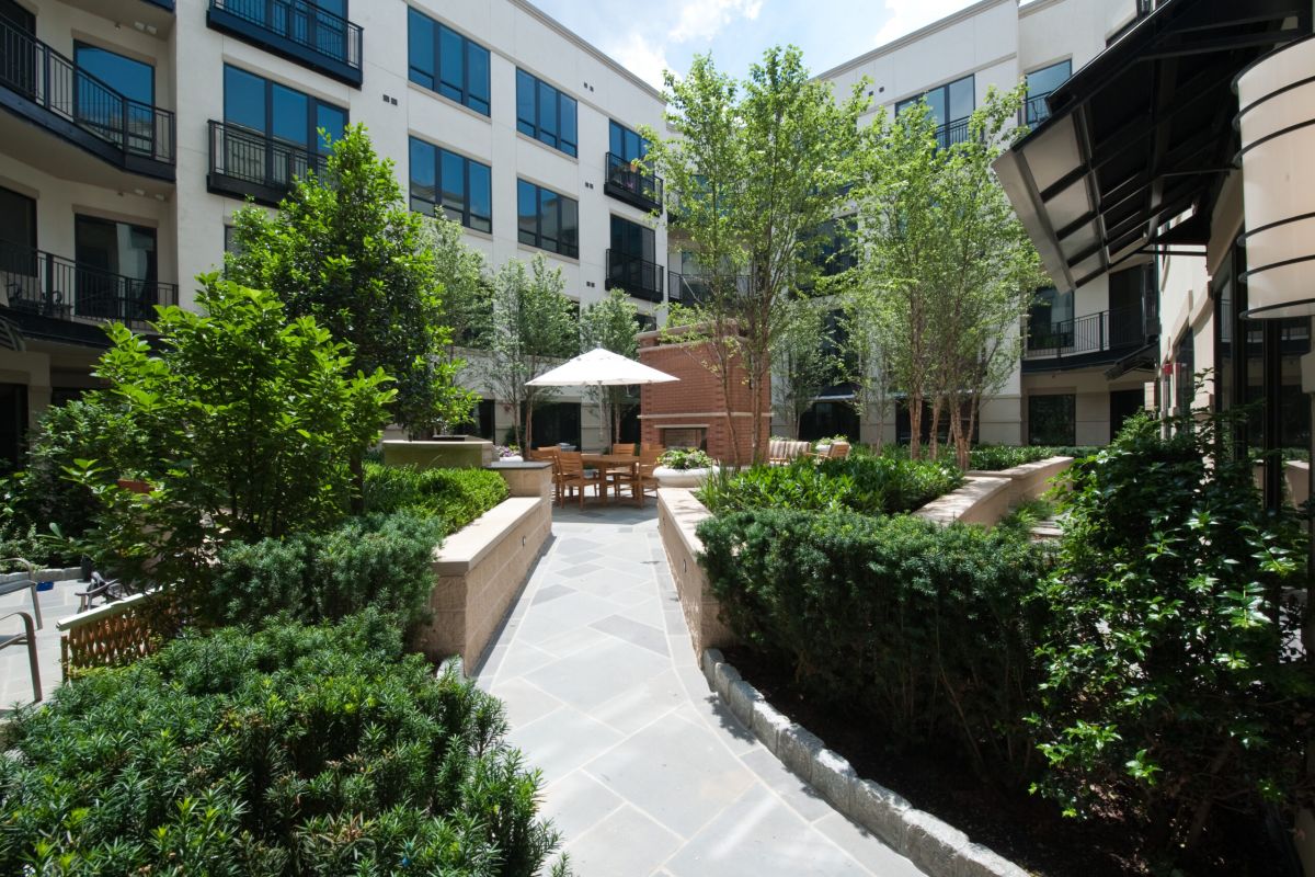 Sunny courtyard with green plants, outdoor seating, and umbrella surrounded by modern apartment buildings.