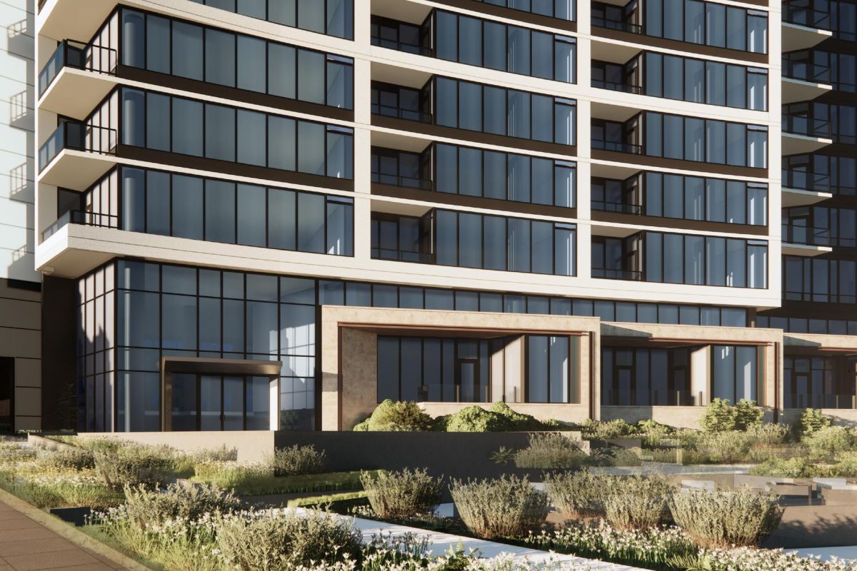 Modern glass building with balconies and landscaped area in the foreground, under a clear blue sky.