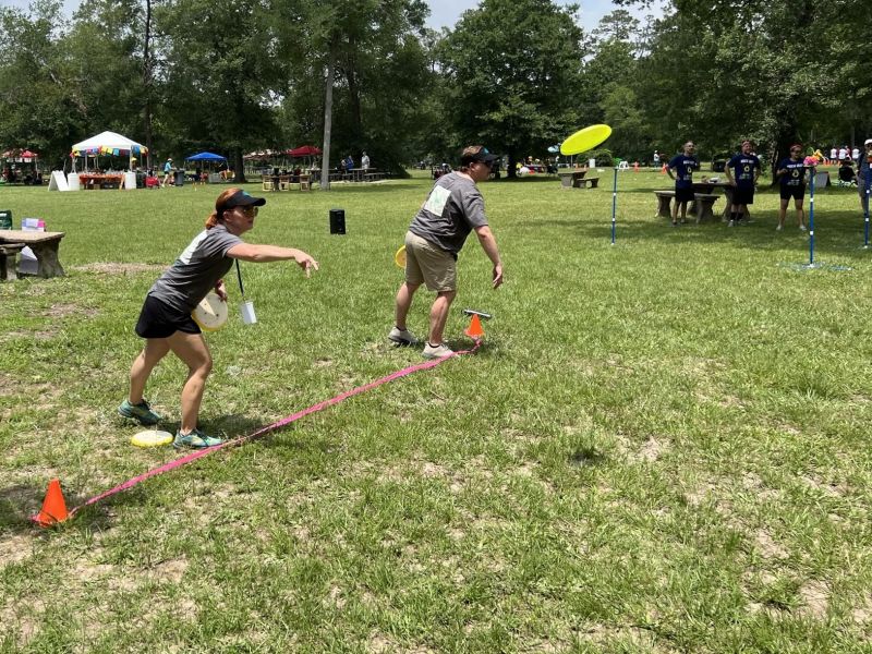 Two people throw frisbees toward targets at an outdoor event with spectators and tents in the background.
