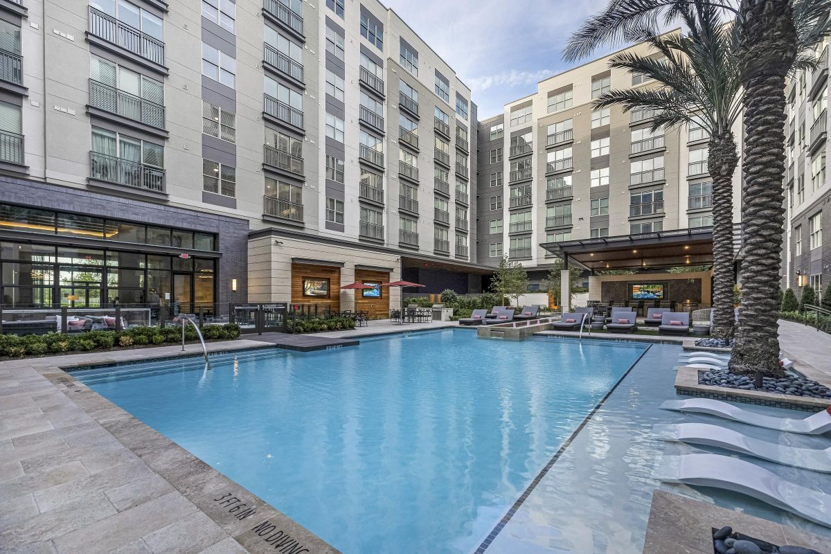 Modern apartment courtyard with a pool, lounge chairs, and palm trees, surrounded by multi-story buildings.