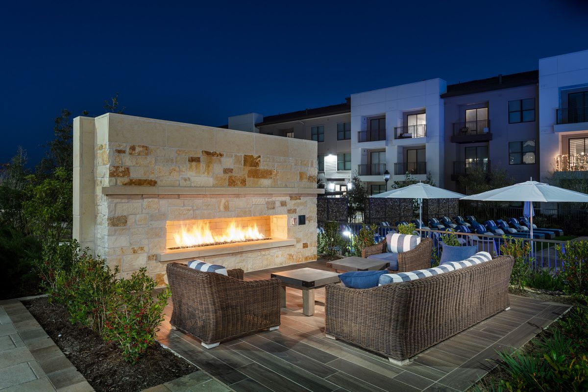 Wicker patio seating by an outdoor fireplace at night, with apartments and pool area in the background.