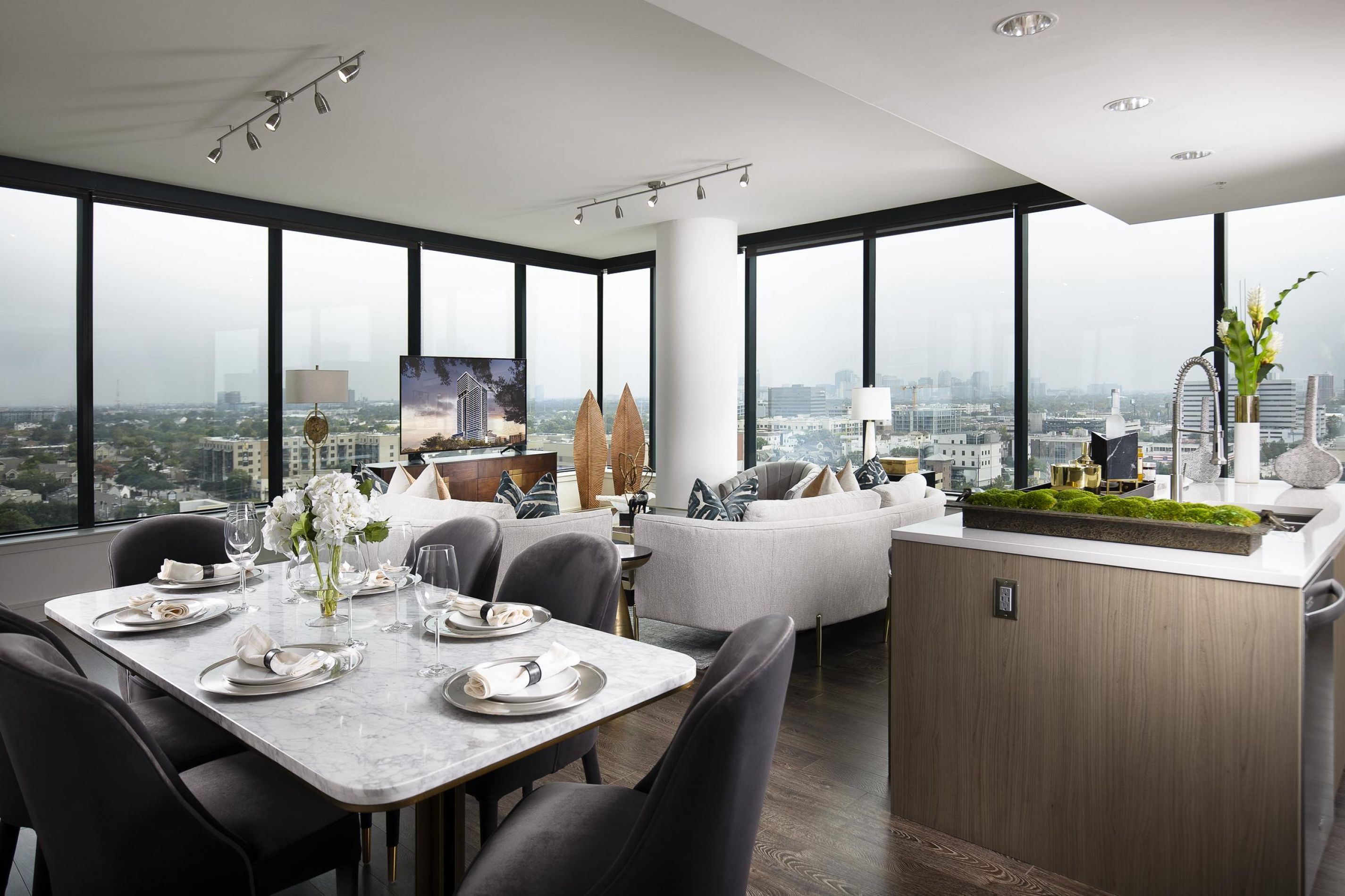 Modern open-plan apartment with dining and living areas, large windows, city views, gray furnishings, and a kitchen island.