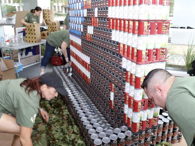 Three people build a large structure out of canned goods indoors, arranging cans in rows and patterns.