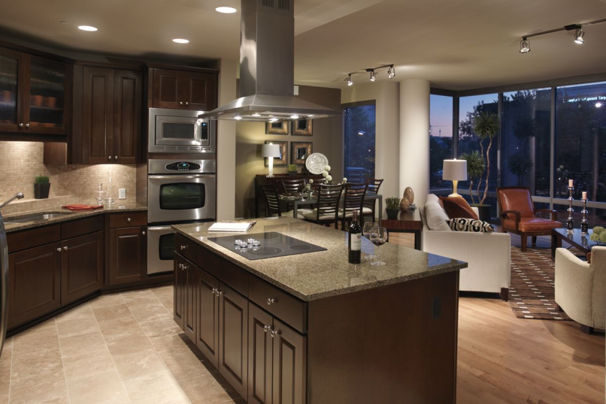Modern kitchen with dark wood cabinets, granite island, and open living room with large windows at dusk.