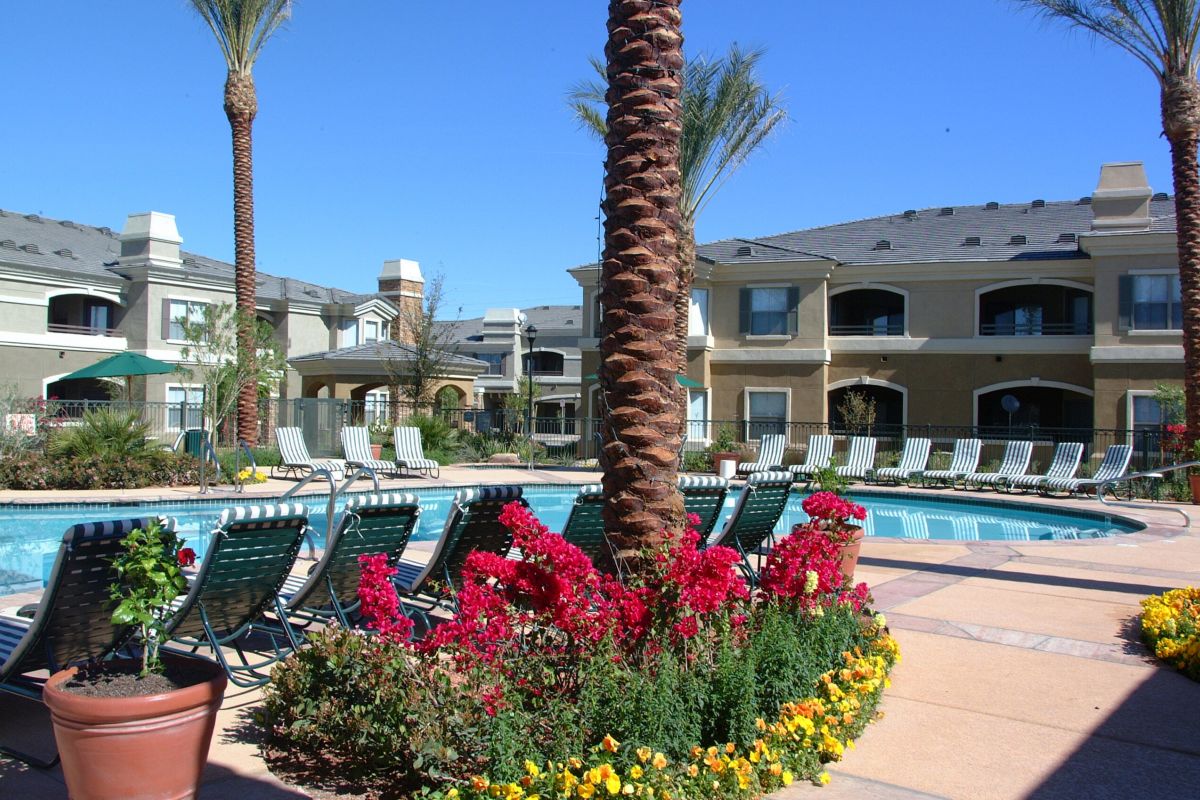 Apartment pool area with lounge chairs, palm trees, and colorful flowers on a sunny day.