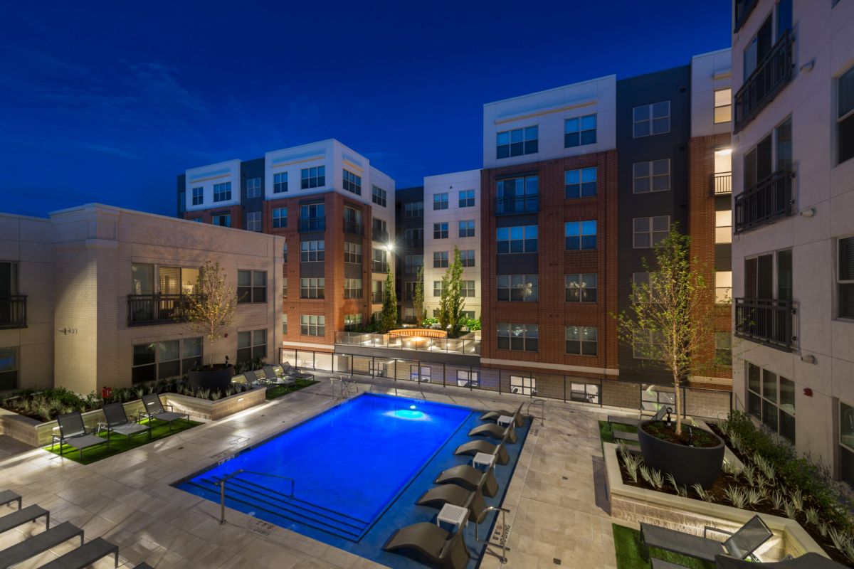Night view of a modern apartment complex with a lit outdoor pool and lounge chairs around it.