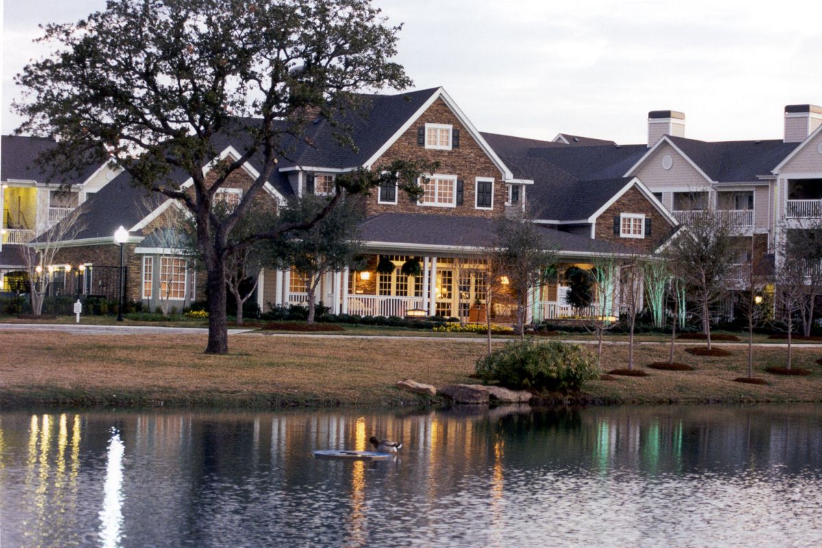 Large house with a porch by a pond, trees, and a duck swimming in the water at dusk.
