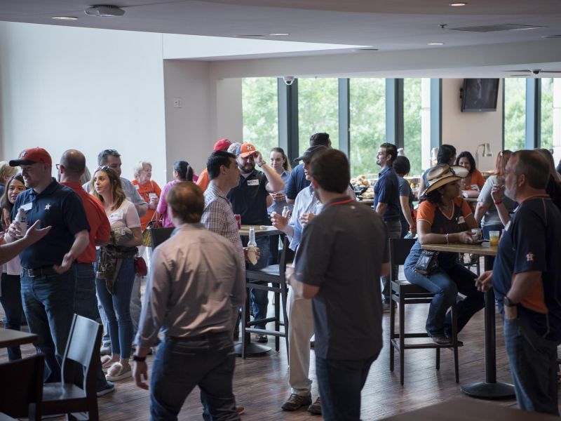 A group of people socialize and mingle in a bright indoor space with large windows and high tables.