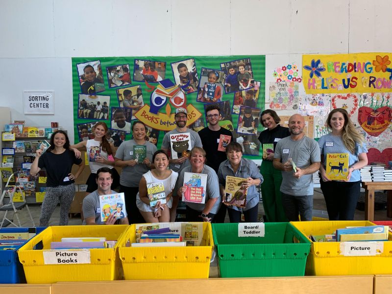 A group of volunteers smile while holding children’s books in a colorful reading center with bins of books.