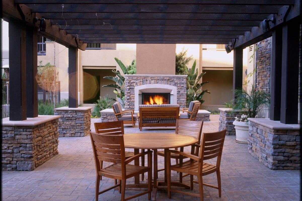 Outdoor patio with wooden table and chairs, stone fireplace, and pergola with plants in the background.