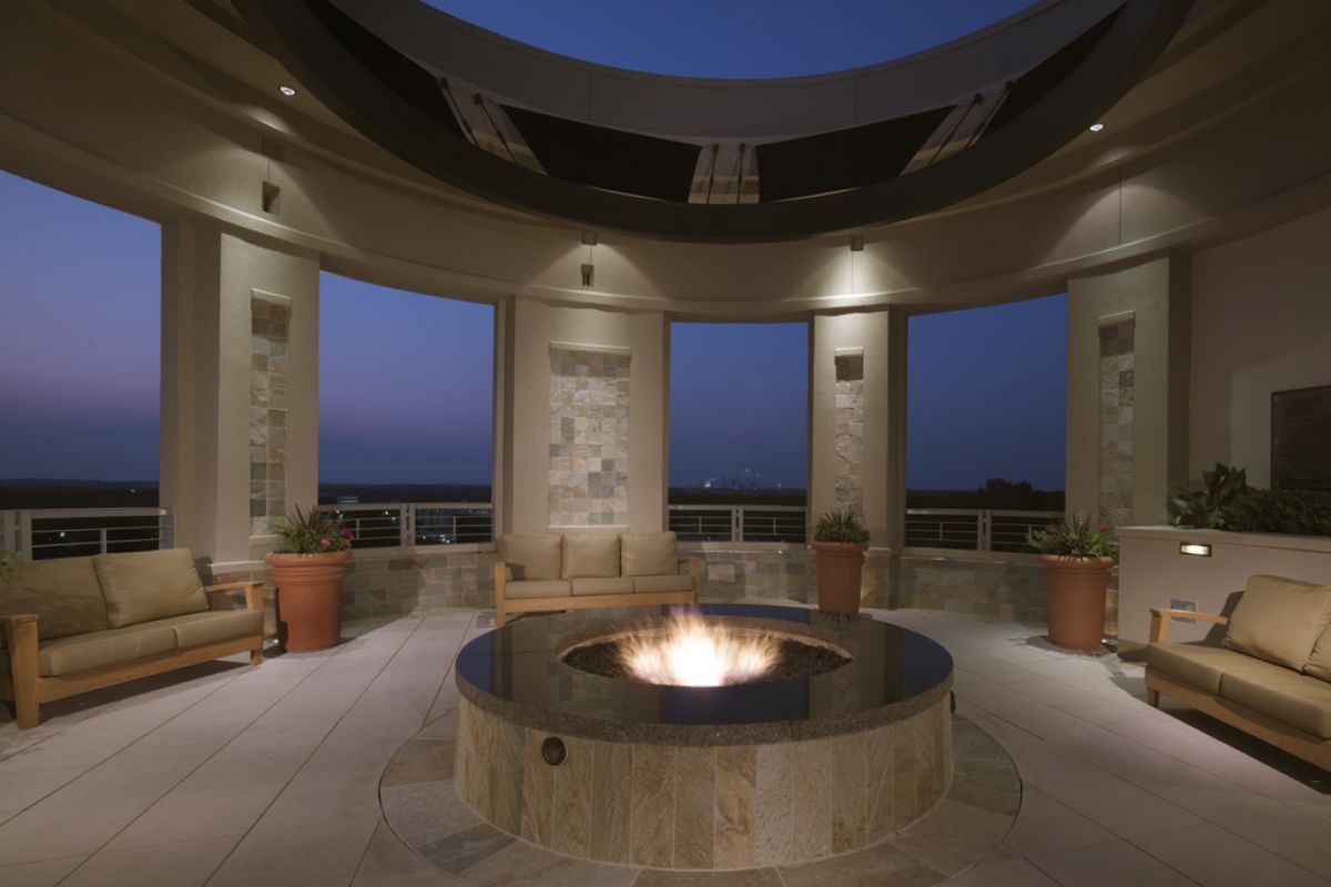 Modern outdoor lounge with a circular fire pit, couches, and potted plants under a domed open roof at dusk.