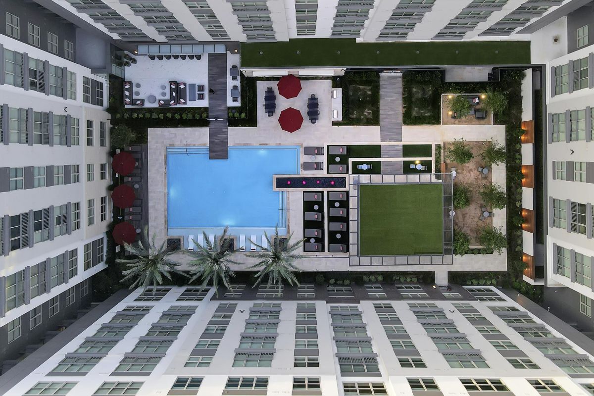 Aerial view of a modern building courtyard with a pool, seating areas, palm trees, and green lawn.