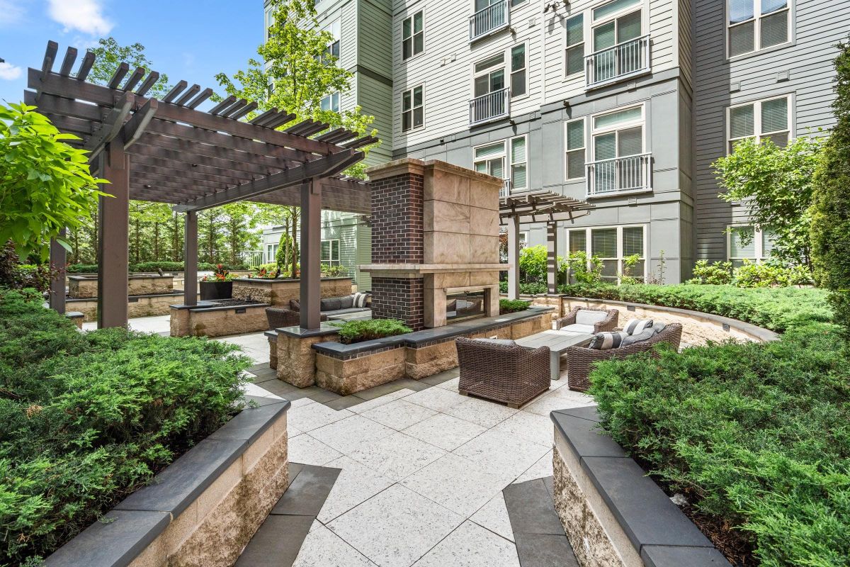 Outdoor patio with modern seating, fireplace, and pergolas, surrounded by greenery and tall building.