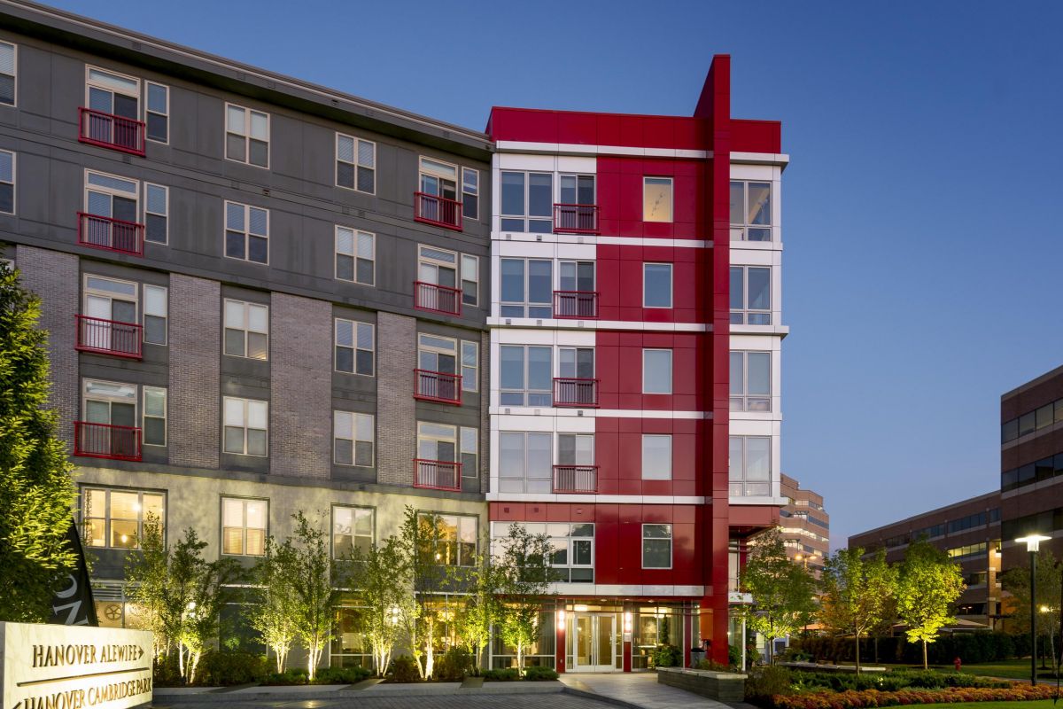 Modern apartment building with red accents, surrounded by trees, at dusk. Sign reads "Hanover Alewife.