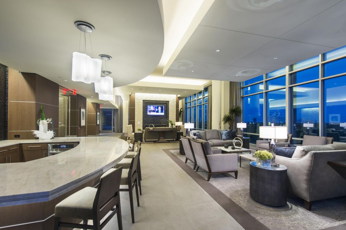 Modern lounge with curved bar, sofas, and large windows. Neutral tones and ambient lighting create a cozy atmosphere.